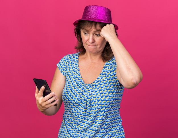 Donna di mezza età con cappello da festa che guarda lo schermo del suo smartphone confusa mentre celebra la festa di compleanno in piedi sul muro rosa