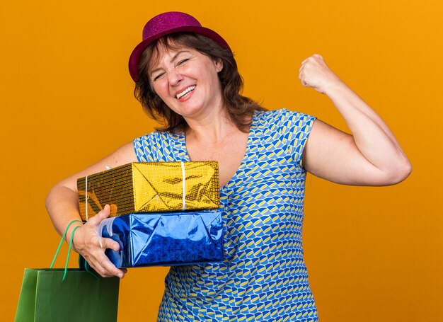 Donna di mezza età con cappello da festa che tiene in mano un sacchetto di carta con regali di compleanno felice ed eccitato che stringe il pugno