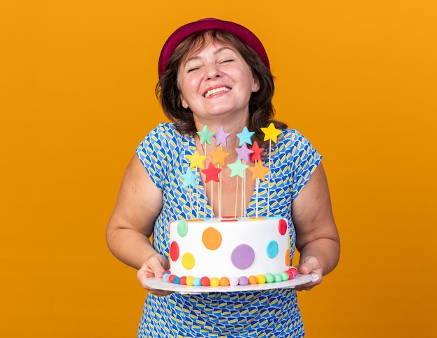 Donna di mezza età con cappello da festa che tiene in mano una torta di compleanno sorridendo allegramente felice ed emozionata che celebra la festa di compleanno in piedi sul muro arancione