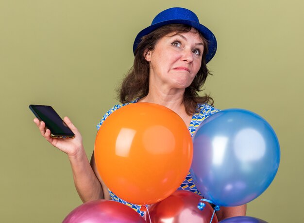 Donna di mezza età con cappello da festa con un mazzo di palloncini colorati che tengono lo smartphone alzando lo sguardo perplesso