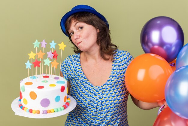 Donna di mezza età con cappello da festa con un mazzo di palloncini colorati che tengono torta di compleanno confusa