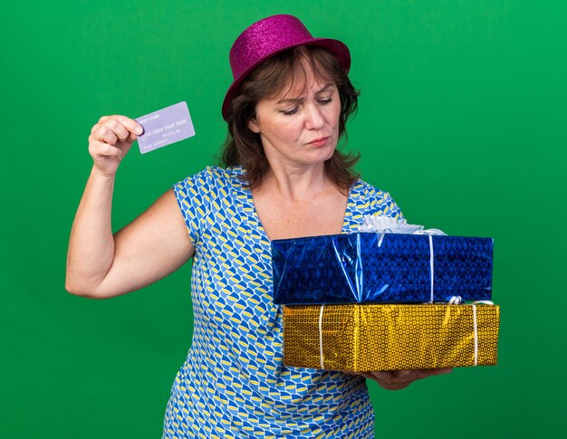 Donna di mezza età con cappello da festa in possesso di regali di compleanno e carta di credito che sembra confusa mentre celebra la festa di compleanno in piedi sul muro verde