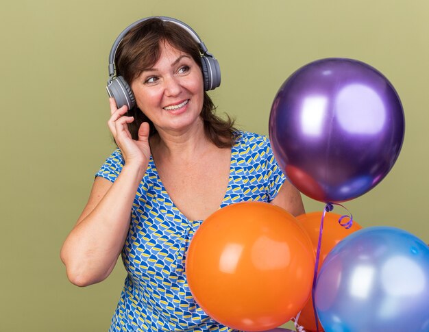 Donna di mezza età con le cuffie e un mucchio di palloncini colorati felice e allegra che si gode la sua musica preferita