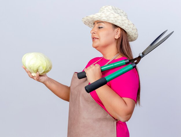 Donna di mezza età del giardiniere in grembiule e cappello che tiene cavolo e tagliasiepi guardando il cavolo con un sorriso sul viso