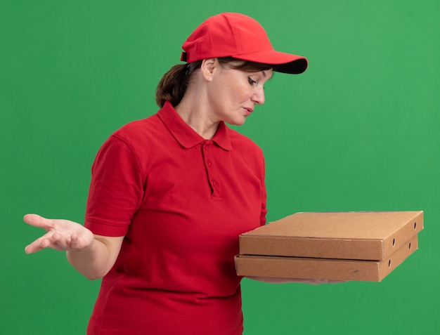 Donna di mezza età delle consegne in uniforme rossa e cappuccio che tiene le scatole della pizza guardandole confuse in piedi sopra il muro verde