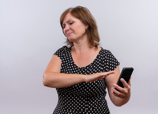 Donna di mezza età dispiaciuta che tiene il telefono cellulare e che indica con la mano su fondo bianco isolato