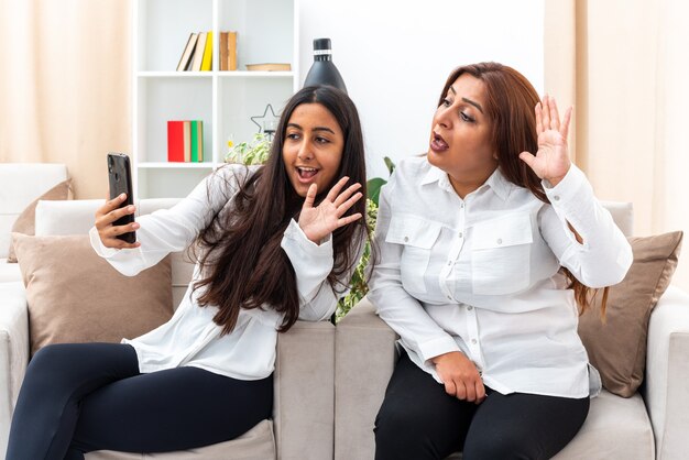 Donna di mezza età e la sua giovane figlia in camicie bianche e pantaloni neri seduti sulle sedie con smartphone con videochiamata felice e positiva che saluta con le mani in soggiorno luminoso