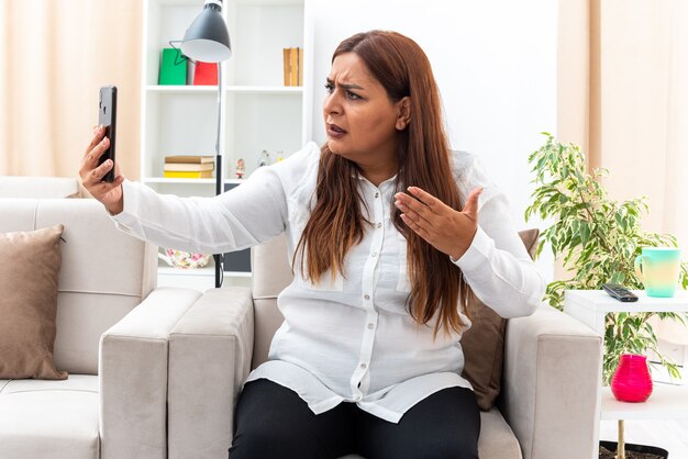 Donna di mezza età in camicia bianca e pantaloni neri che ha videochiamata utilizzando smartphone cercando confuso e scontento seduto sulla sedia in soggiorno luminoso