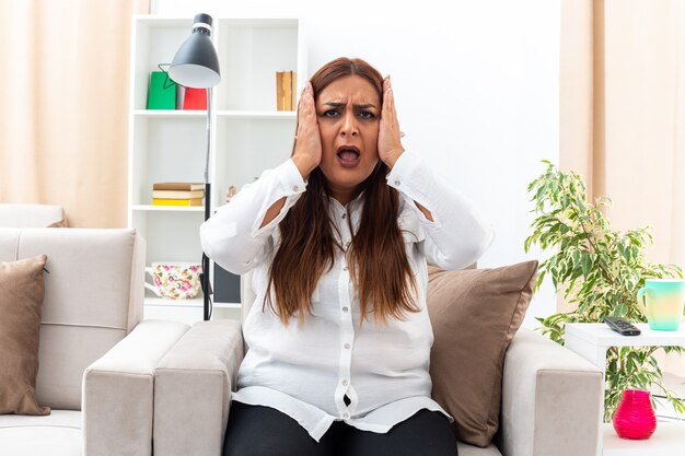 Donna di mezza età in camicia bianca e pantaloni neri frustrata con le mani sulla testa seduta sulla sedia in un soggiorno luminoso