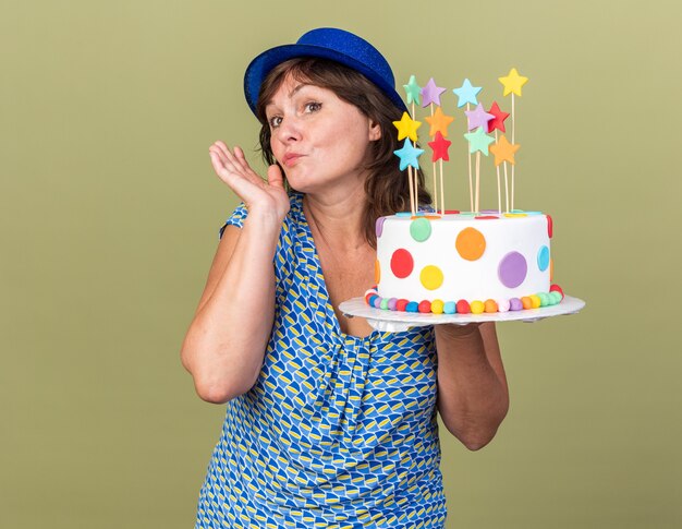 Donna di mezza età in cappello da festa che tiene la torta di compleanno che guarda da parte con un sorriso timido sul viso che celebra la festa di compleanno in piedi sopra il muro verde