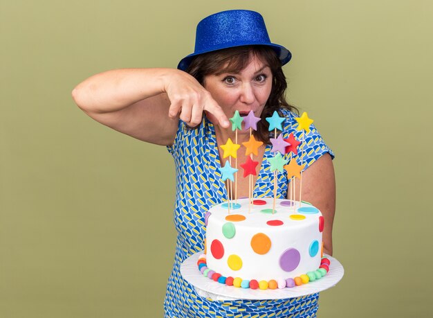 Donna di mezza età in cappello da festa che tiene la torta di compleanno che indica con il dito indice felice e sorpresa che celebra la festa di compleanno in piedi sul muro verde green