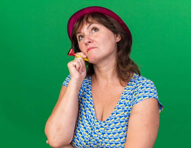 Donna di mezza età in cappello da festa con fischietto alzando lo sguardo perplesso che celebra la festa di compleanno in piedi sul muro verde