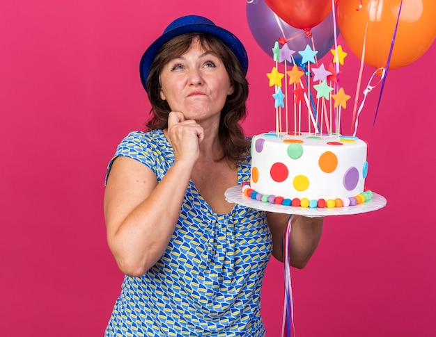 Donna di mezza età in cappello da festa con palloncini colorati che tengono la torta di compleanno alzando lo sguardo con espressione pensosa pensando di celebrare la festa di compleanno in piedi sul muro rosa