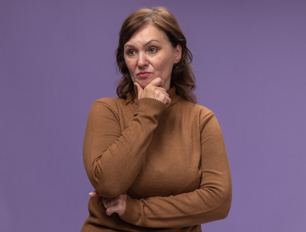 Donna di mezza età in dolcevita marrone che osserva da parte con la faccia seria con la mano sul mento pensando in piedi sopra il muro viola