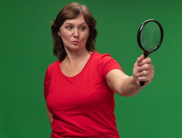 Donna di mezza età in maglietta rossa che tiene la lente d'ingrandimento guardandolo con la faccia seria che sta sopra la parete verde