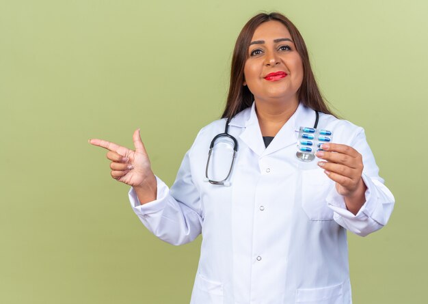 Donna di mezza età medico in camice bianco con stetoscopio che tiene blister con pillole sorridendo allegramente puntando con il dito indice sul lato in piedi sul verde