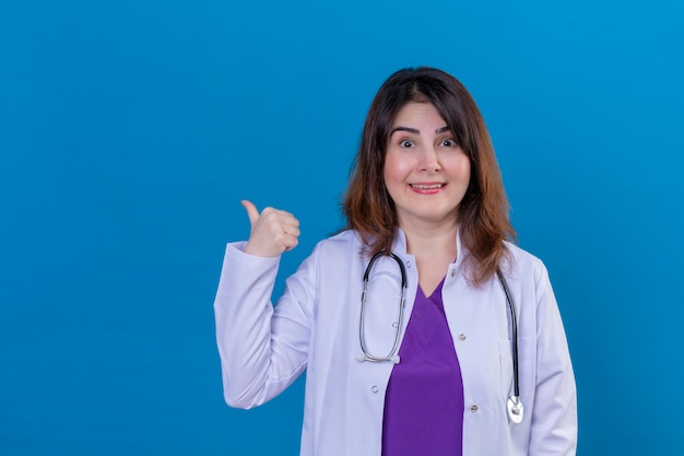 Donna di mezza età medico indossa camice bianco e con uno stetoscopio guardando la telecamera sorridendo allegramente indicando il lato con il pollice in alto in piedi su sfondo blu
