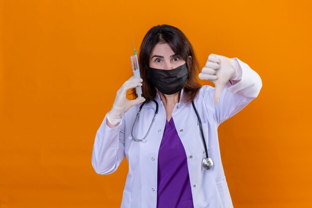 Donna di mezza età medico indossa camice bianco nella maschera facciale protettiva nera e con uno stetoscopio tenendo la siringa guardando la telecamera che mostra i pollici verso il basso in piedi su sfondo arancione