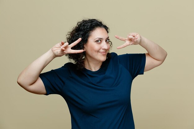 Donna di mezza età soddisfatta che indossa una maglietta che mostra il segno di pace con entrambe le mani vicino agli occhi guardando la fotocamera isolata su sfondo verde oliva