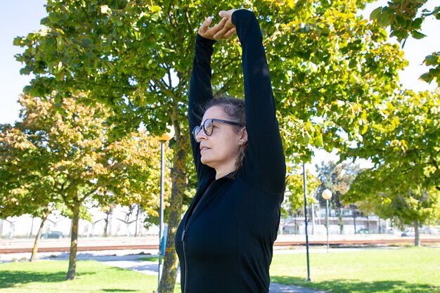 Donna di mezza età sportiva focalizzata che allunga il corpo, alzando le mani, distogliendo lo sguardo mentre si esercita nel parco. Benessere o concetto di stile di vita attivo