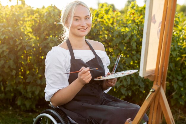 Donna di smiley in sedia a rotelle all'aperto nella natura con tavolozza e tela