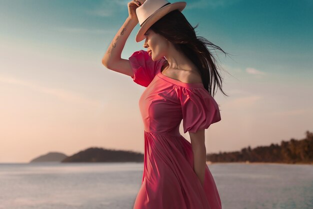 Donna di stile estivo all'aperto, ritratto di moda soleggiata di donna sensuale indossa un abito rosa sul tramonto della spiaggia in riva al mare