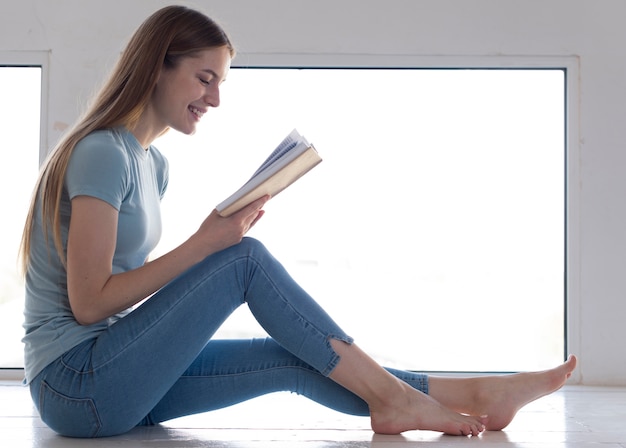 Donna di vista laterale che legge un libro accanto alla finestra