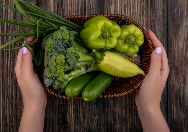 Donna di vista superiore che tiene i peperoni verdi con i broccoli dei cetrioli e nel cestino delle cipolle verdi su fondo di legno