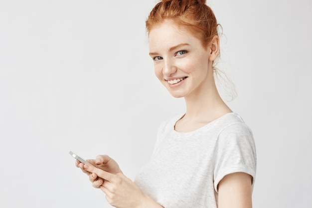 donna di zenzero sorridente azienda telefono twitting o utilizzando i social media sul muro bianco