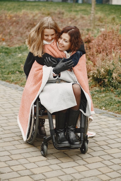 Donna disabile in sedia a rotelle con la figlia. Famiglia che cammina fuori al parco. La bambina ha coperto sua madre dal plaid.