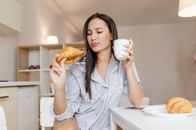 Donna divertente con capelli neri dritti che mangia croissant durante la colazione in appartamenti accoglienti