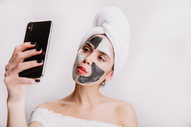 Donna divertente con maschera all'argilla per la cura del viso e in un asciugamano sulla sua testa fa selfie sul muro bianco.