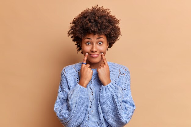 donna divertente fa sorridere con le dita si costringe ad avere un'espressione positiva indossa pose maglione casual contro il muro marrone