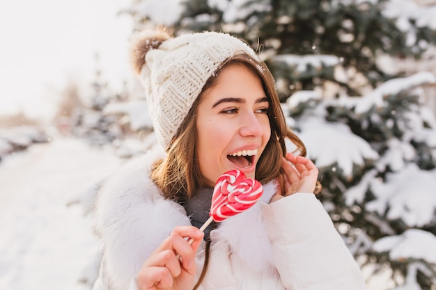 Donna divertente sveglia del ritratto del primo piano in cappello di lana bianco divertendosi con il lecca-lecca cuore rosa sulla strada. Bella giovane donna che gode del freddo inverno, neve, emozioni luminose.