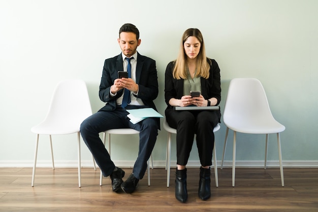 Donna e uomo d'affari professionisti che guardano i loro smartphone e mandano messaggi mentre aspettano un colloquio di lavoro mentre sono seduti in un ufficio di reclutamento