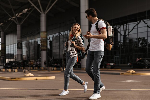 Donna e uomo gioiosi in t-shirt e jeans bianchi camminano e parlano vicino all'aeroporto Ritratto a figura intera di viaggiatori con zaini