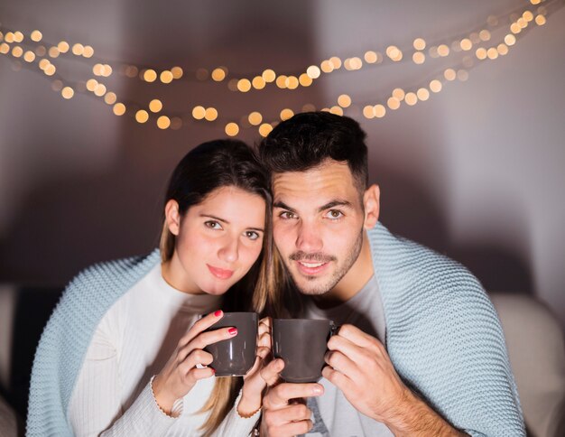 Donna e uomo in coverlet con tazze sul divano in camera oscura