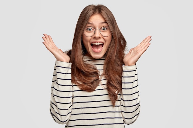 Donna eccitata con espressioni facciali stupite, tenere le mani vicino al viso, vestita con abiti a righe casual, modello contro il muro bianco. La foto della donna allegra diffonde le palme, ha un'espressione felice