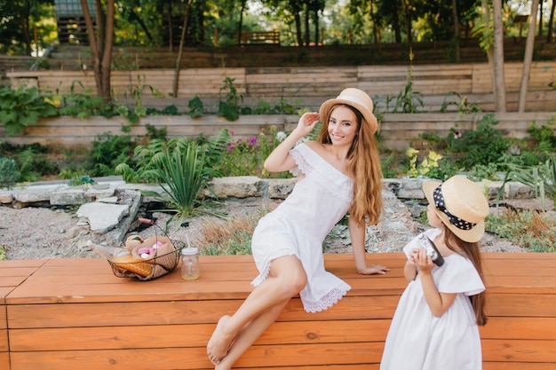 Donna eccitata in cappello vintage in posa sulla natura mentre sua figlia la guarda con interesse. Ritratto all'aperto dal retro della bambina in abito bianco in piedi accanto a un'aiuola con la mamma.