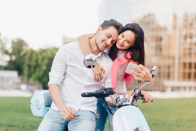 Donna elegante che tocca delicatamente l'uomo bello sullo scooter e che fa selfie con lui