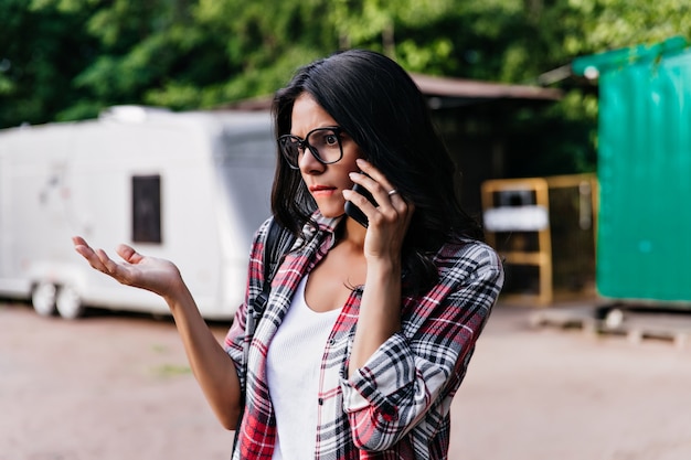 Donna elegante dispiaciuta in occhiali alla moda parlando al telefono la mattina. Colpo esterno della ragazza attraente in posa con l'espressione del viso triste sulla strada.