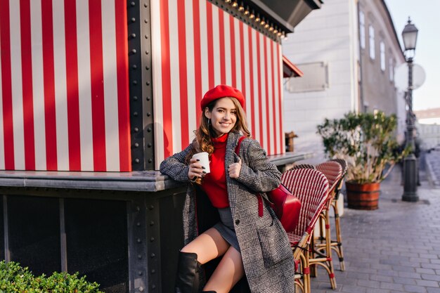 Donna elegante in gonna e cappotto che beve caffè per strada con un sorriso dolce