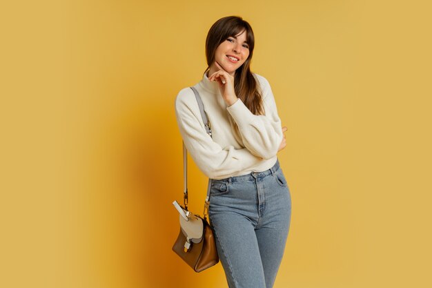 Donna elegante in posa in studio su giallo. Indossa un maglione di lana bianca e jeans.