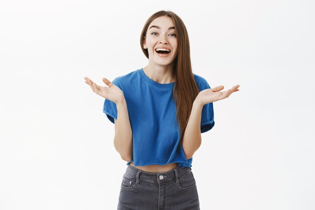 Donna emotiva alla moda intrattenuta sorpresa con lunghi capelli castani in maglietta blu ritagliata che applaude le mani dal divertimento e dalla felicità sorridendo con gioia e guardando entusiasta oltre il muro grigio