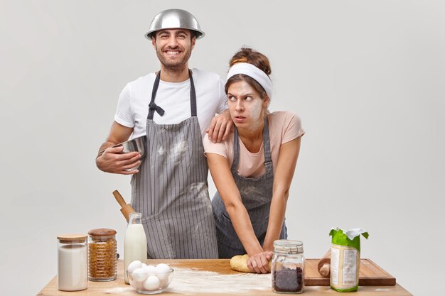 Donna esausta con farina sul viso, impasta la pasta, stanca di preparare il pane fatto in casa, uomo allegro in grembiule si appoggia sulla spalla, tiene una ciotola, felice di aiutare la moglie in cucina. Le coppie fanno la pizza insieme