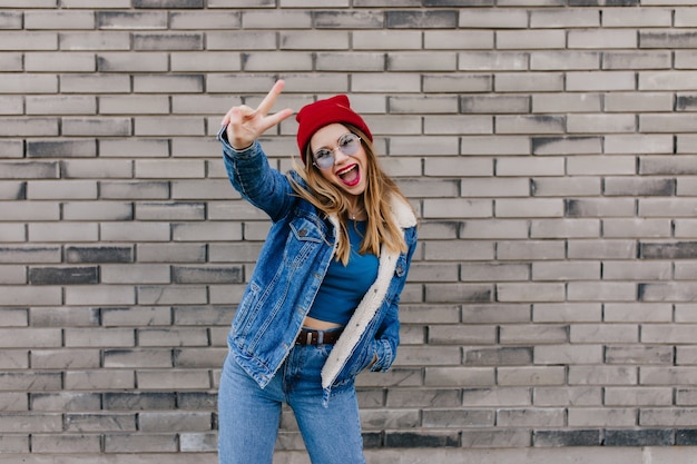 Donna europea eccitata con espressione del viso felice ballando sul muro di mattoni. Colpo esterno della ragazza alla moda positiva in cappello rosso scherzare sulla strada.