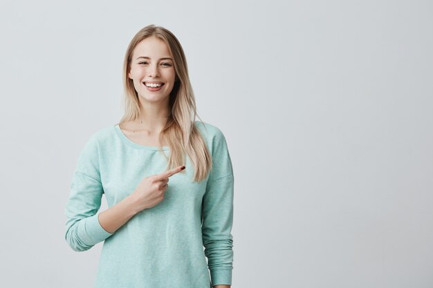 Donna europea positiva allegra sorridente che porta camicia blu-chiaro che indica il suo dito indice da parte allo spazio della copia