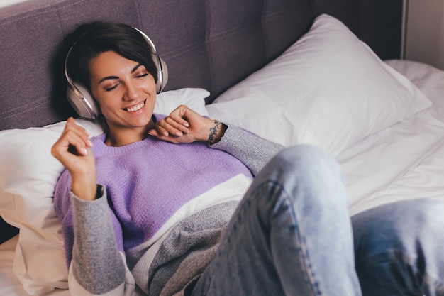 Donna felice a casa sul letto comodo indossando pullover vestiti caldi, ascoltare musica