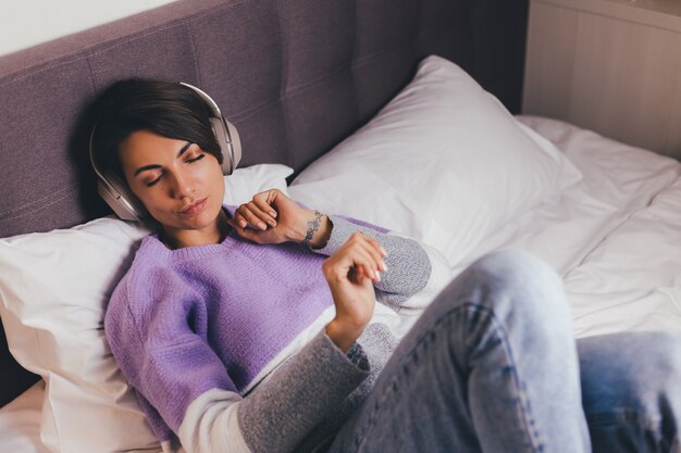 Donna felice a casa sul letto comodo indossando pullover vestiti caldi, ascoltare musica