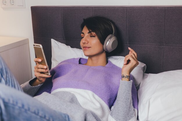 Donna felice a casa sul letto comodo indossando pullover vestiti caldi, ascoltare musica